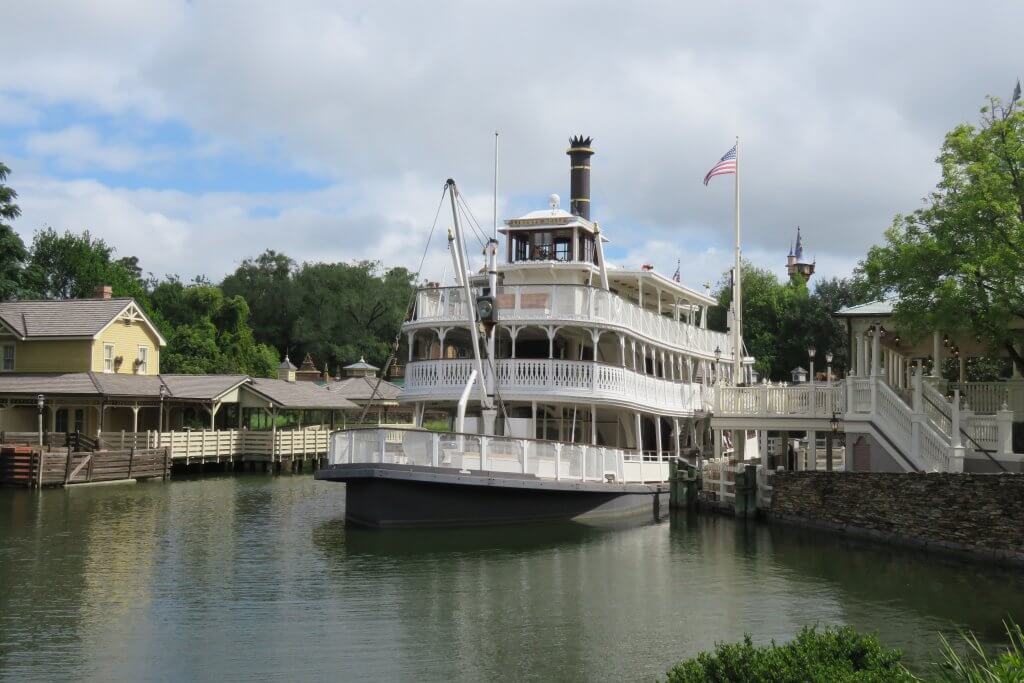 The Liberty Belle riverboat docked along the Rivers of America with the American flag flying above and Cinderella Castle spire in the distance