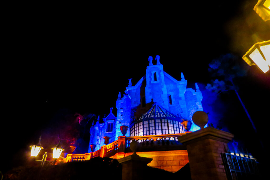 The Haunted Mansion glowing blue at night with lanterns and light fog—spooky Halloween ambiance at Magic Kingdom.