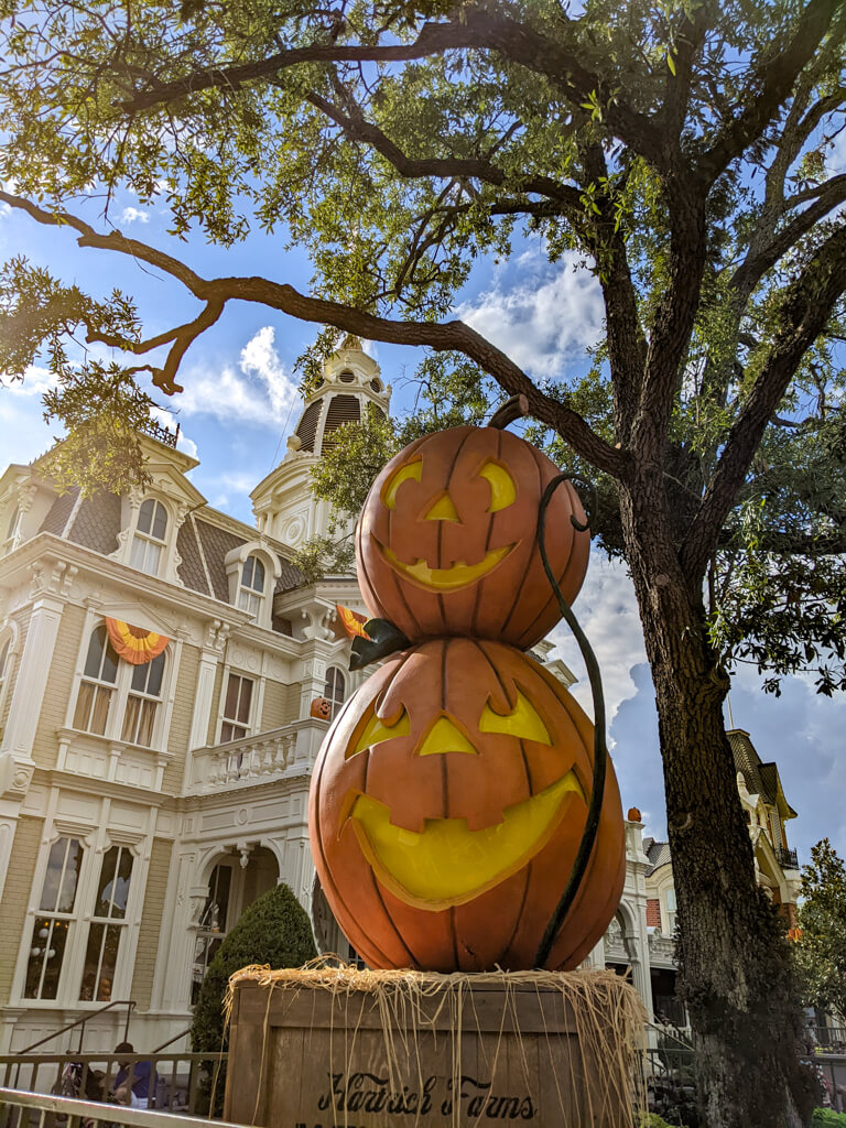 Two stacked jack-o’-lanterns under an oak tree beside Main Street’s Victorian buildings decorated with orange and yellow bunting.
