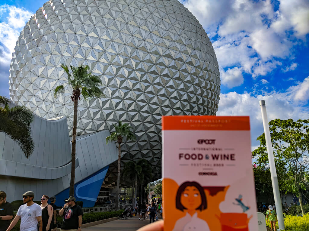 Guest holds the EPCOT International Food & Wine Festival passport in front of Spaceship Earth on a bright fall day.