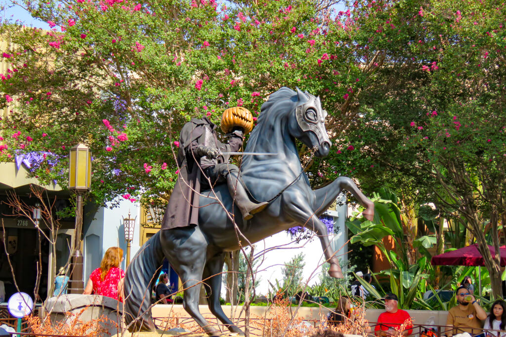 Headless Horseman statue rears on Buena Vista Street—rider holding a jack-o’-lantern and sword amid fall trees at Disney California Adventure.