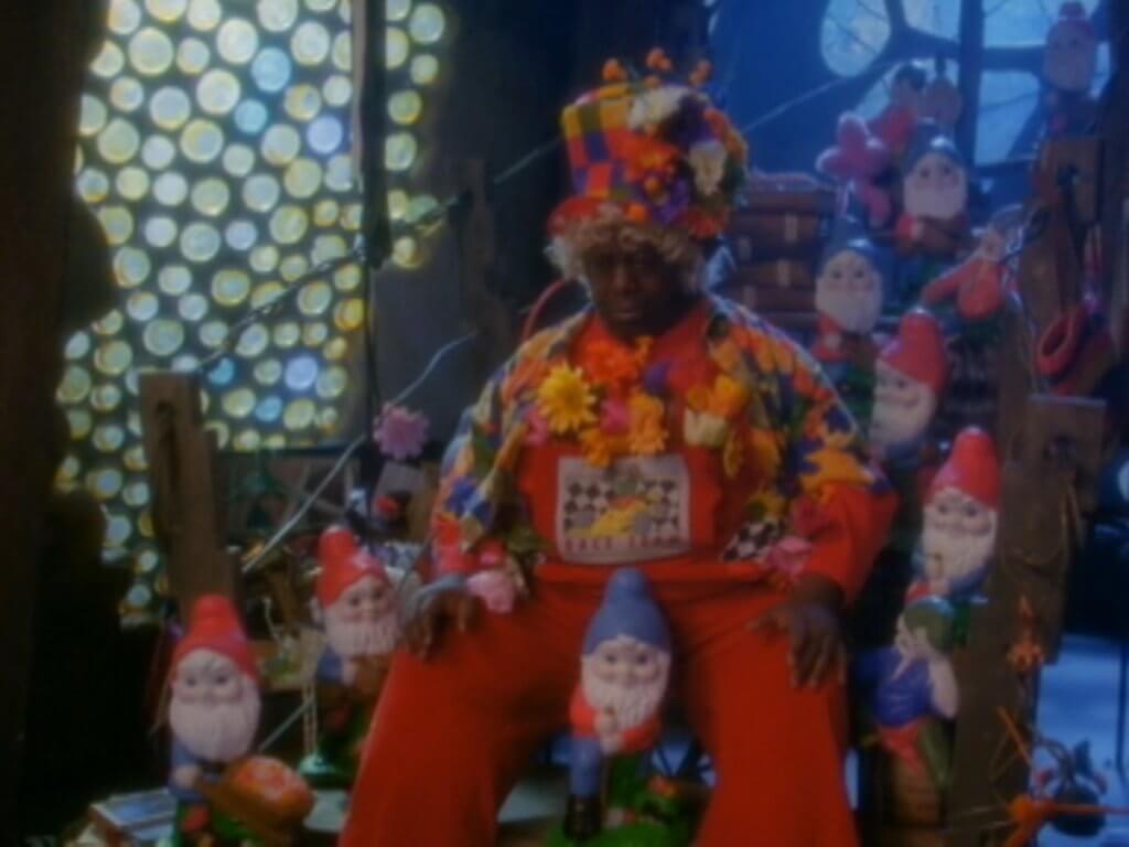 Gort slumped in his cluttered workshop, surrounded by garden gnomes as stained-glass light glows—scene from “Halloweentown II.”