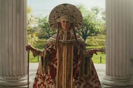 Ornate robed figure in a gilded headdress standing between white columns—dramatic still from Disney’s 2023 “Haunted Mansion.”