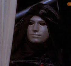 “Phantom of the Megaplex” still—the masked phantom in a dark hood peeks from behind theater curtains, smooth white mask blank and watchful.